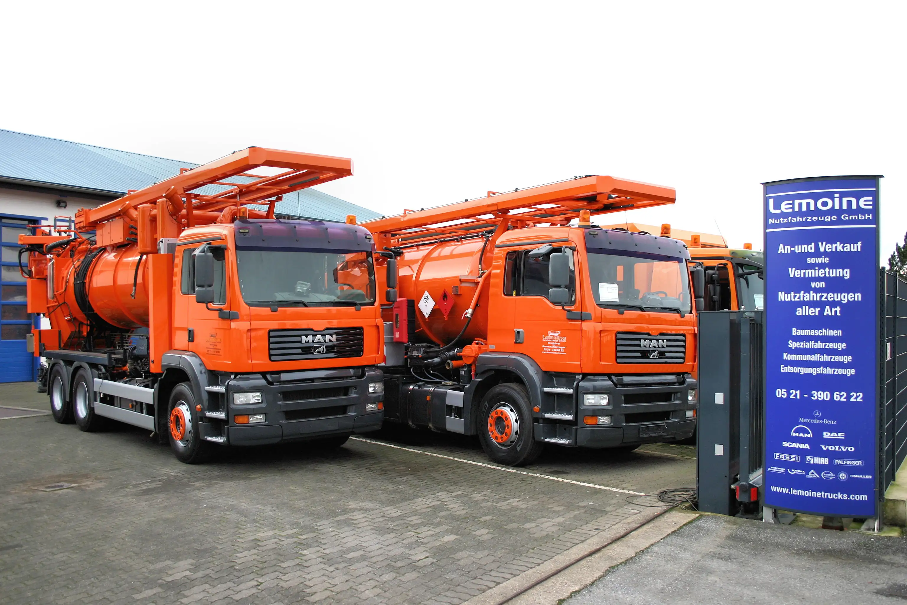 Zwei orange MAN-Saugwagen nebeneinander vor der Werkstatt, rechts das Lemoine-Infoschild